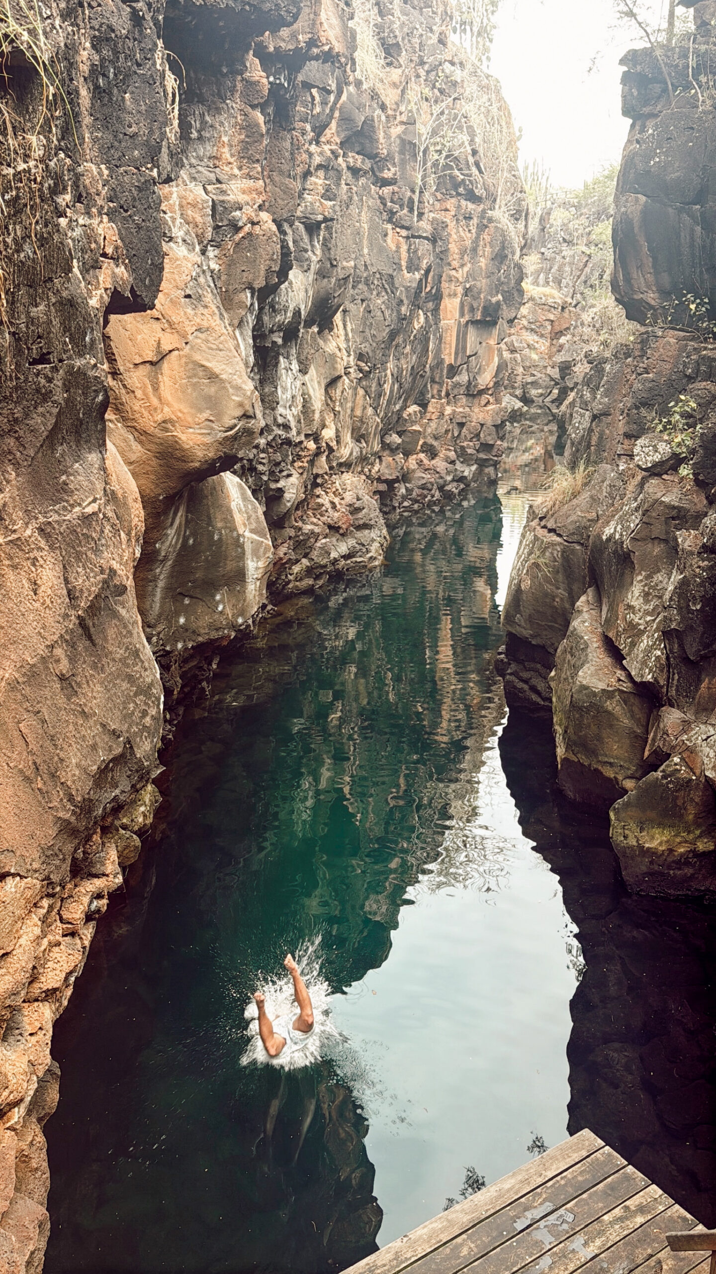 A swimmer diving down into the crystal clear brackish water inside the steep volcanic rock canyon walls.