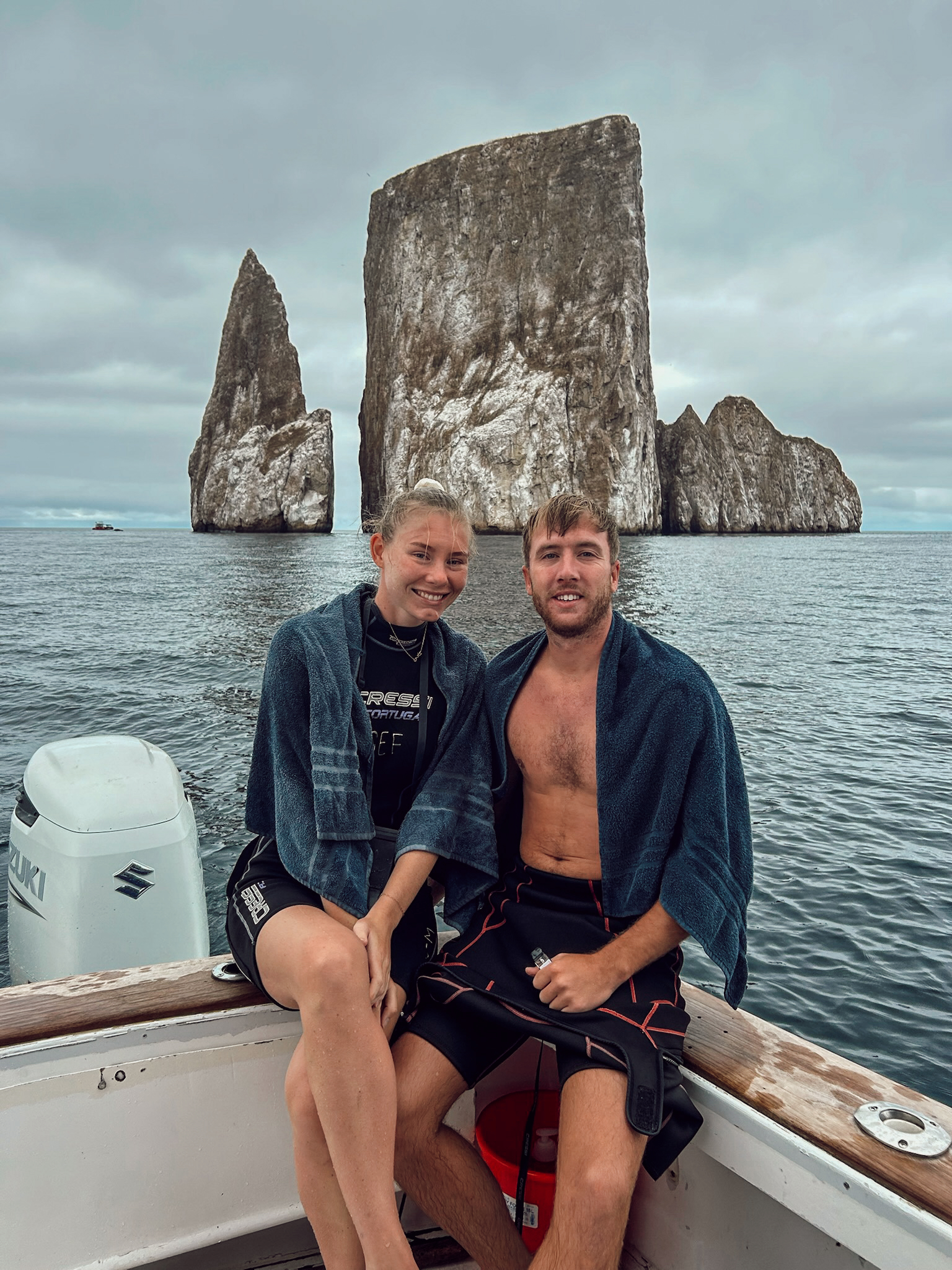 2 travellers smiling on the deck of a boat with the massive dramatic stone walls of Kicker Rock rising out of the sea behind them.