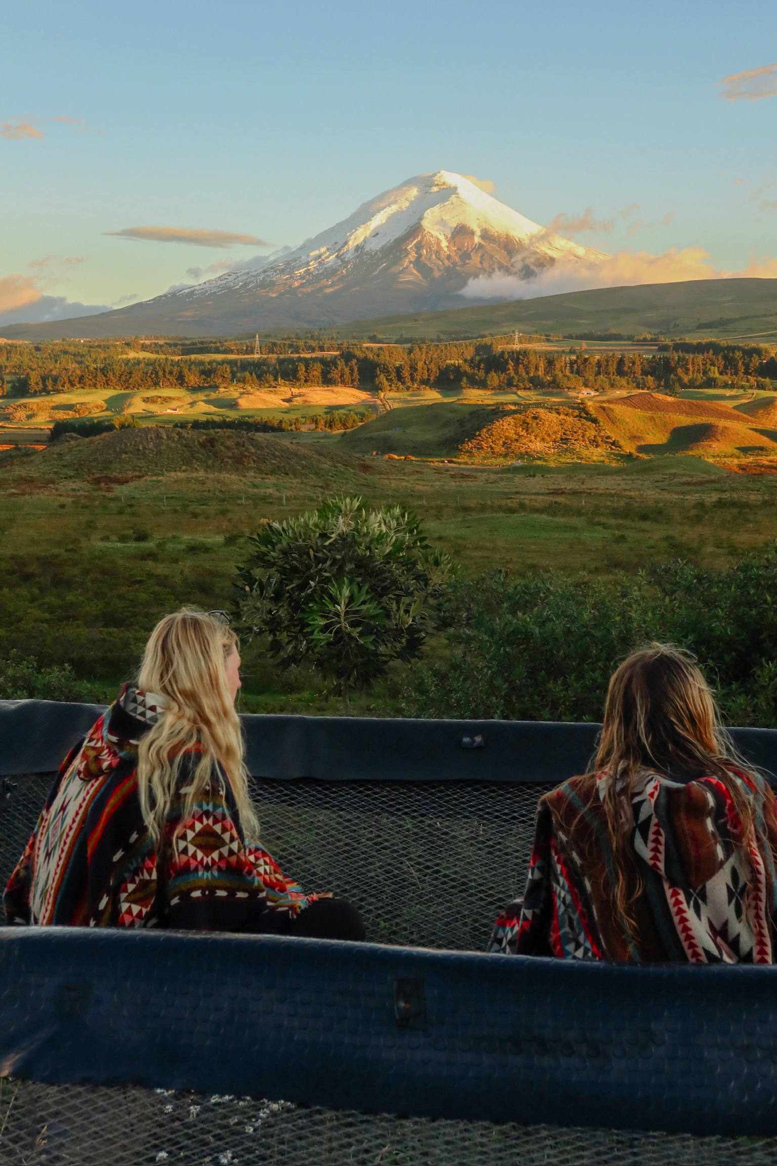 2 girls relaxing and laughing on a giant suspended cliffside netting hammock overlooking the expansive view of the active volcano. Photo taken on the hammocks at the secret garden hostel Cotopaxi in Ecuador 