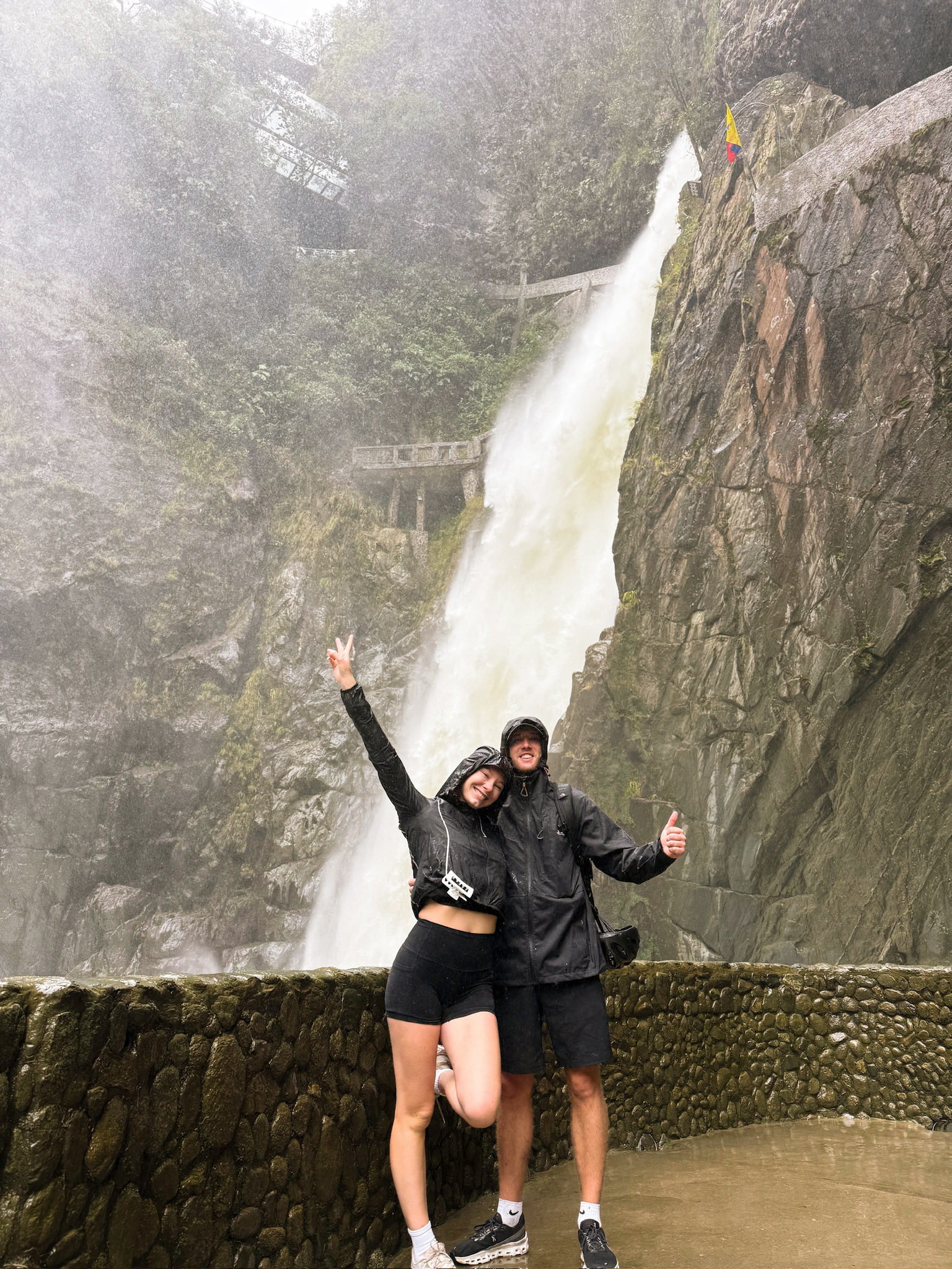 A man and woman smiling and getting completely drenched by water spray on a wooden suspension bridge in front of a massive waterfall.
