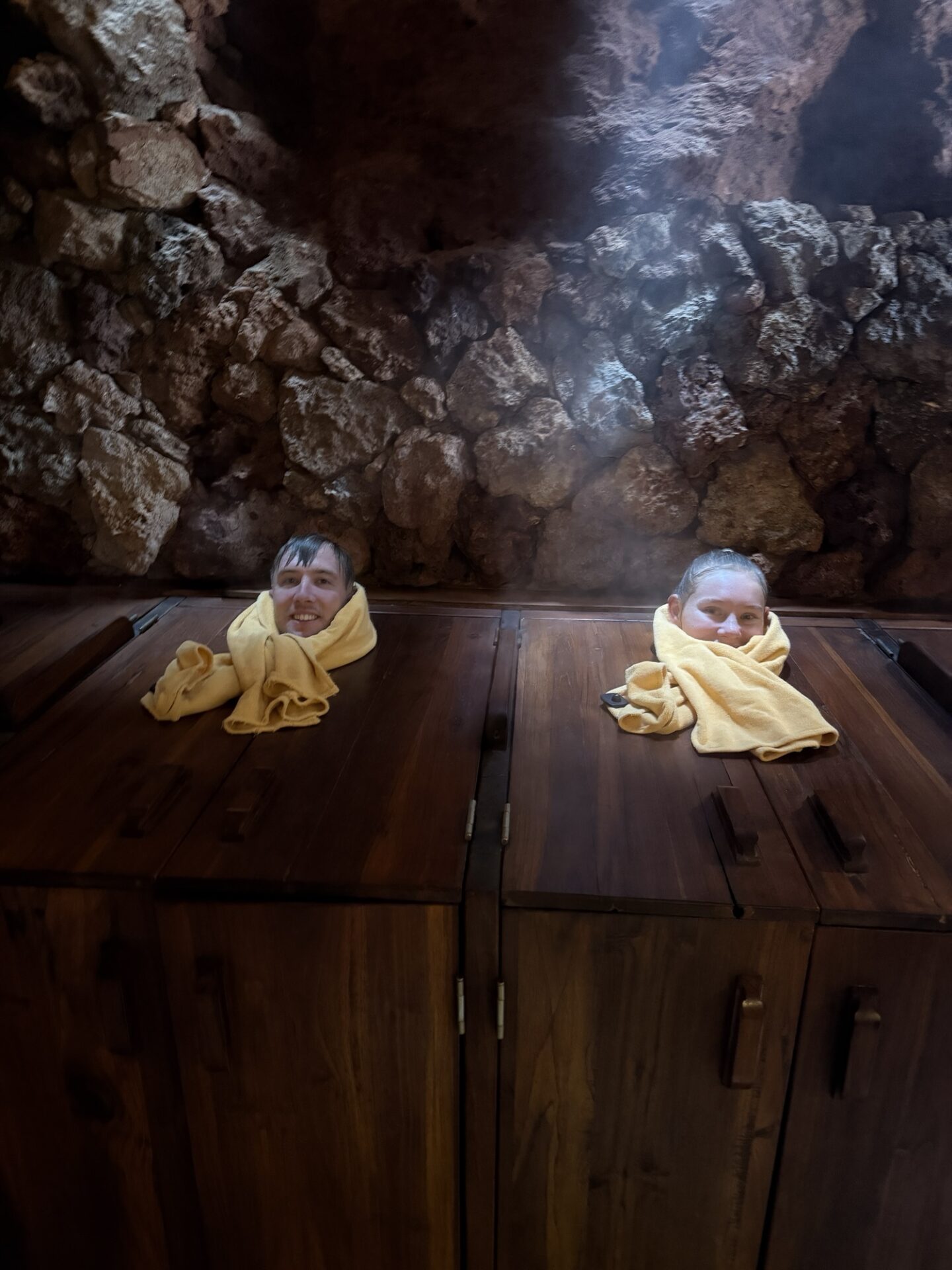 A young couple, Luke and his fiancรฉe, smiling side-by-side with only their heads peeking out of individual wooden volcanic steam boxes at a hot spring spa in Baรฑos de Cuenca, Ecuador.